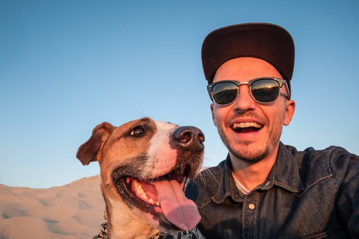 smiling house sitter wearing sunglasses and a cap sits next to a dog who has their mouth open and tongue out
