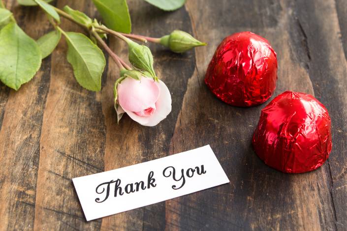 a thank you note from a house sitter on a wooden table alongside some red covered chocolates and a pink rose