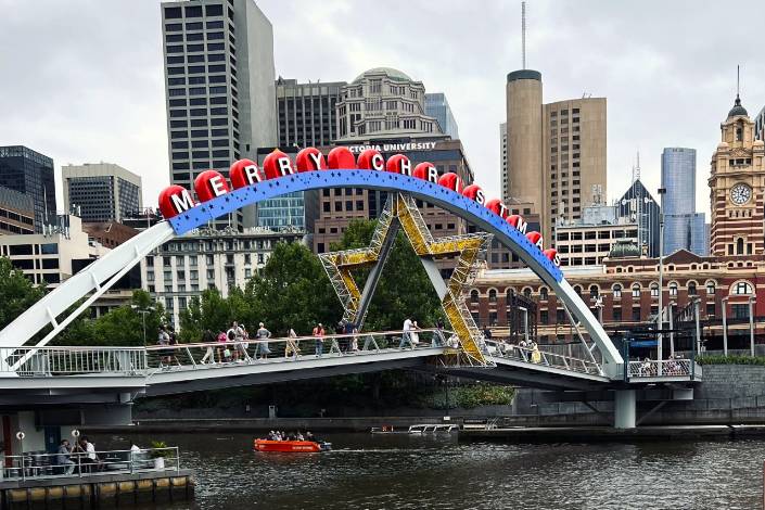 People walking across a bridge with a Merry Christmas sign with building in the background